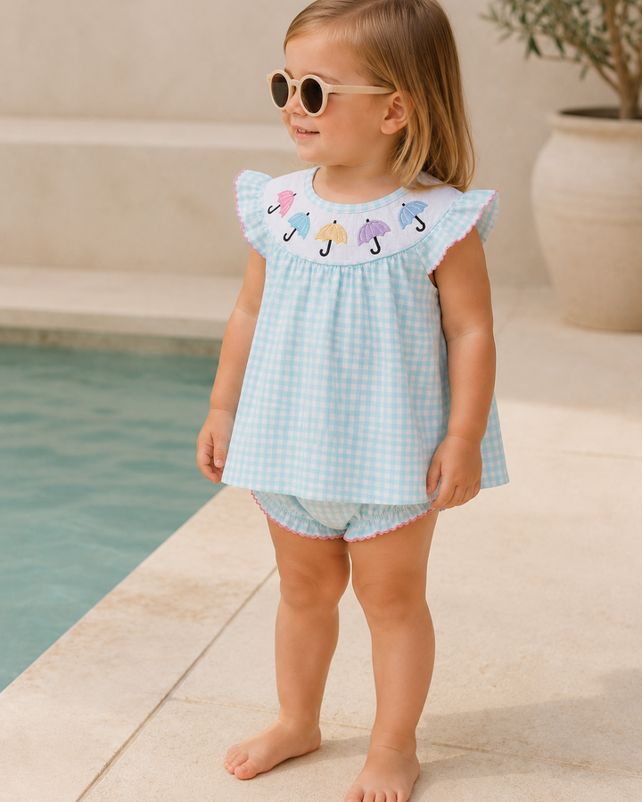 Young girl in a light blue dress with colorful umbrella design standing by a pool.