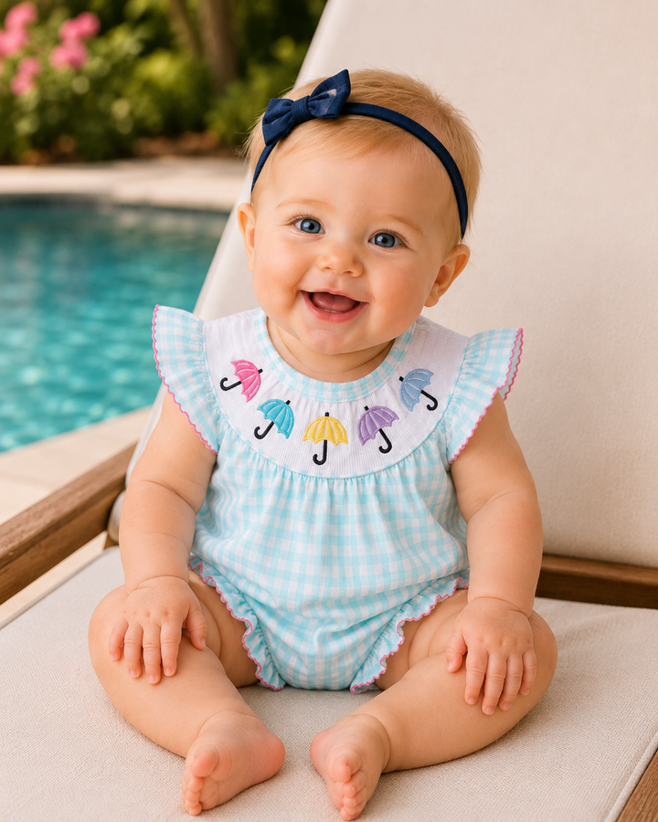 Baby wearing a blue checkered outfit with colorful umbrella design, sitting by a pool.