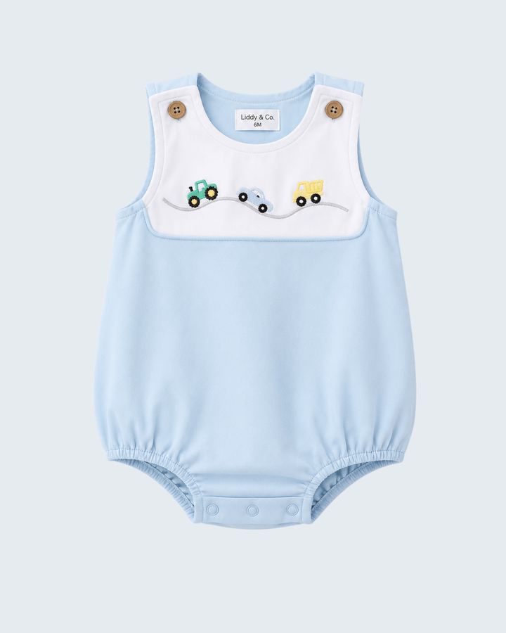 A light blue baby romper with a white bib panel featuring embroidered construction vehicles, photographed flat on a light gray background.
