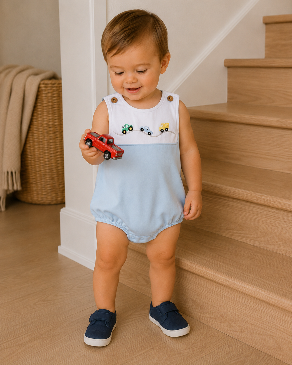 A toddler standing indoors near wooden stairs, wearing a sleeveless light blue romper with a white yoke and small embroidered toy cars, holding a red toy car.