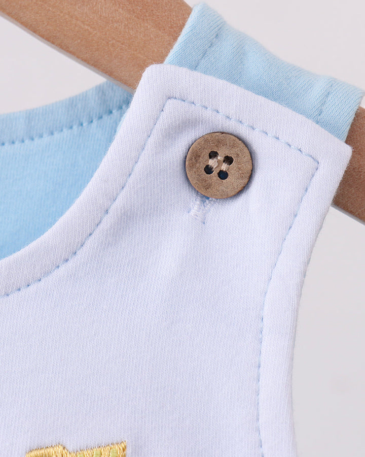 Close-up of a baby garment with a white bib-style panel, light blue inner layer, and a wooden button, shown on a hanger against a light neutral background.