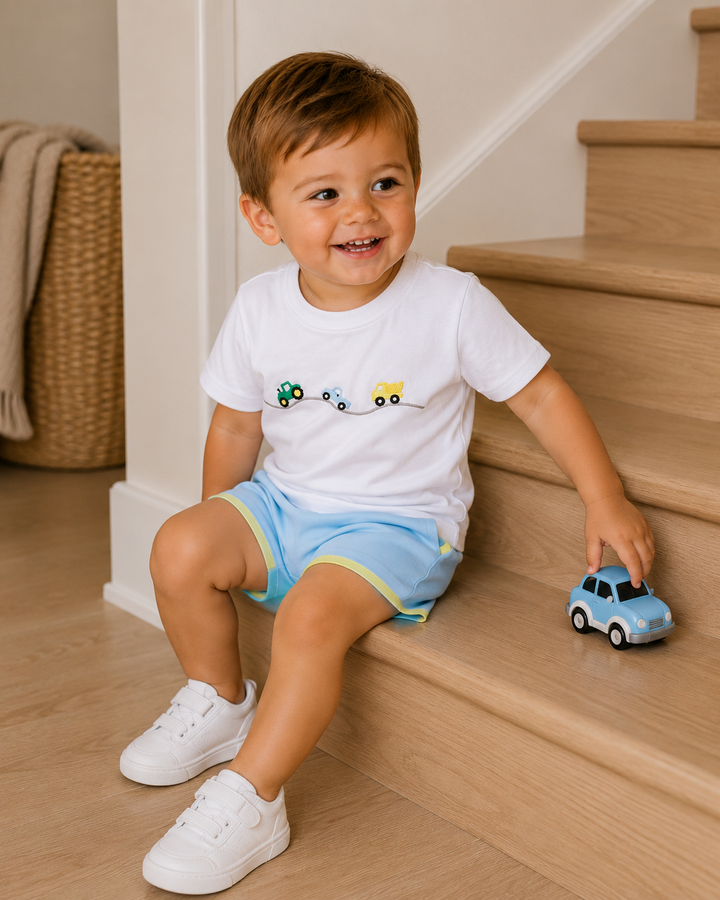 A toddler boy sitting on wooden stairs wearing a white t-shirt with a small road-truck graphic and light blue shorts, holding a small blue toy car.