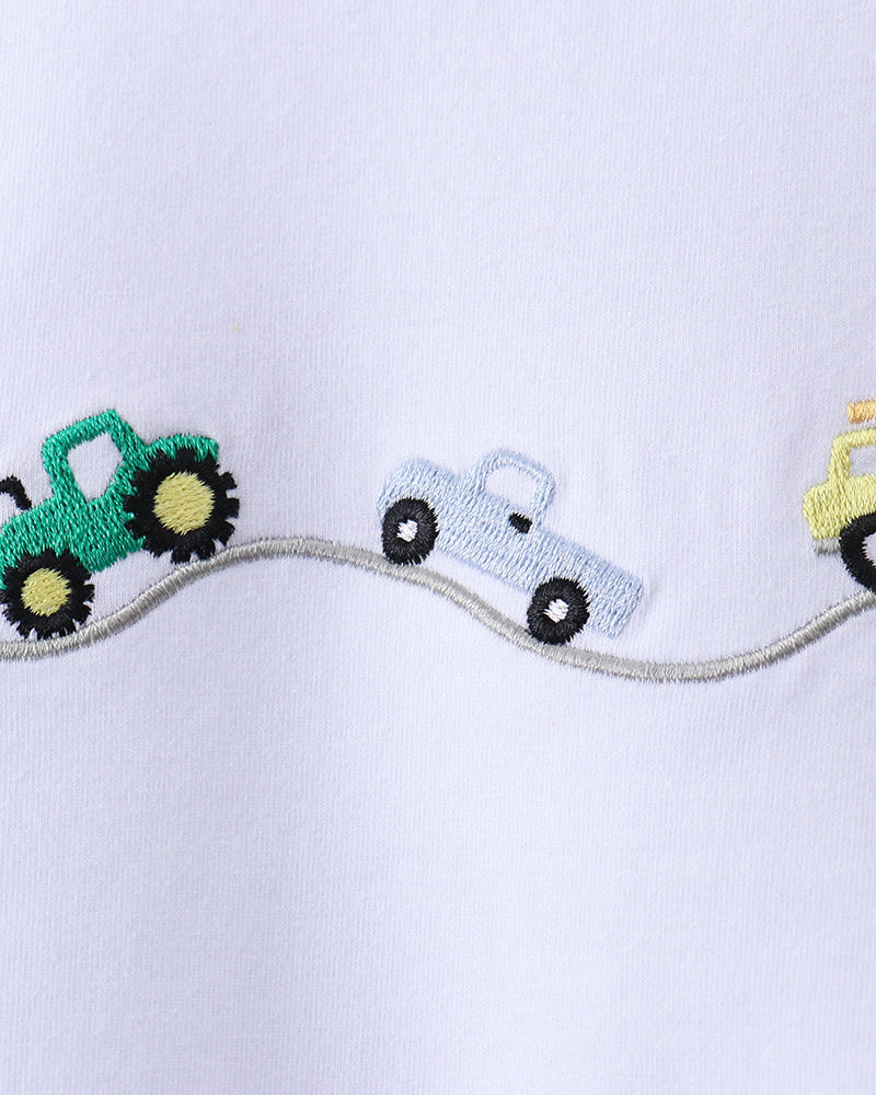 Close-up of a white fabric garment featuring embroidered farm vehicles along a curved gray path, including a green tractor, a light blue pickup truck, and part of a yellow vehicle.