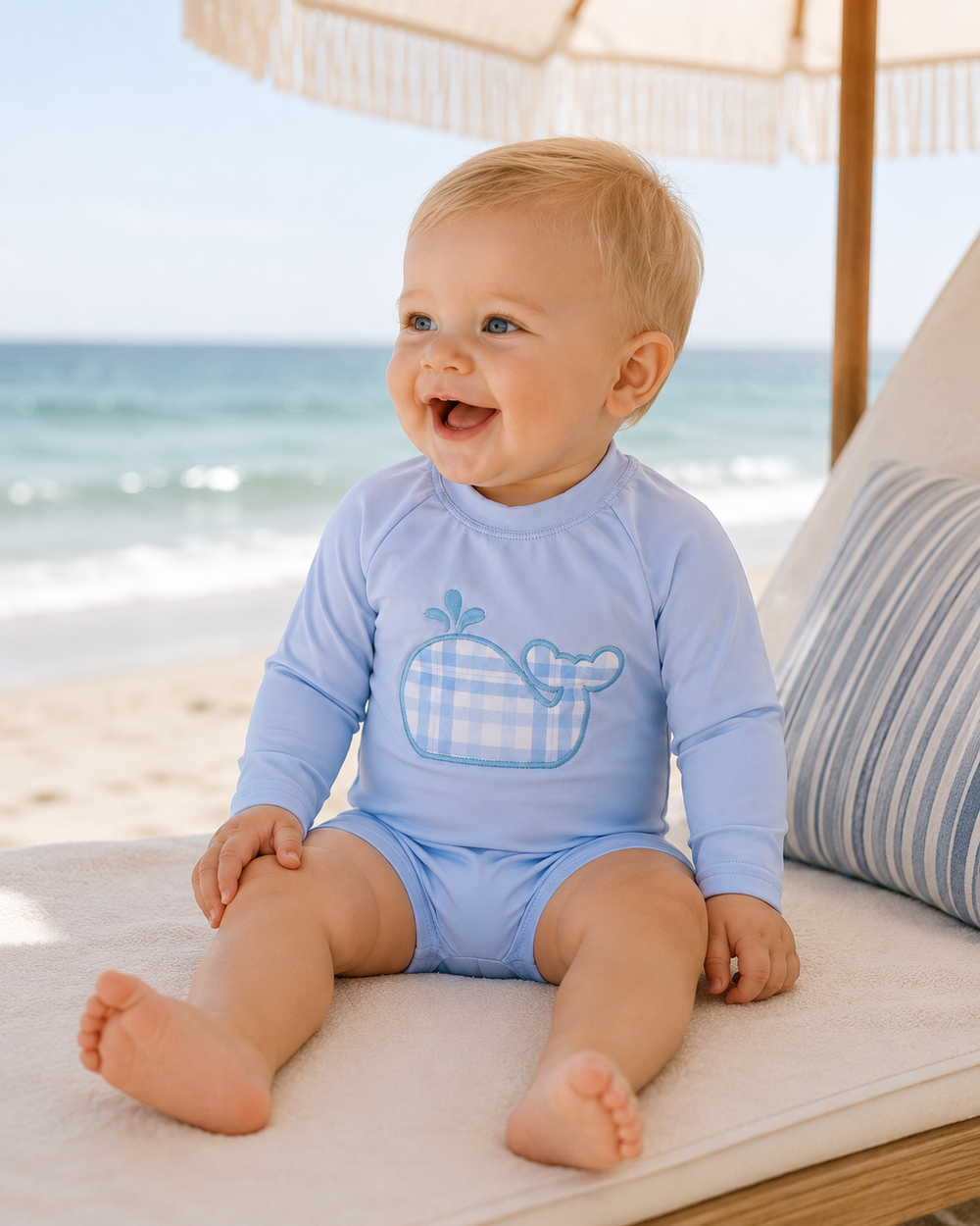 Baby sitting on a beach chair by the ocean wearing a blue swimsuit with a whale design.