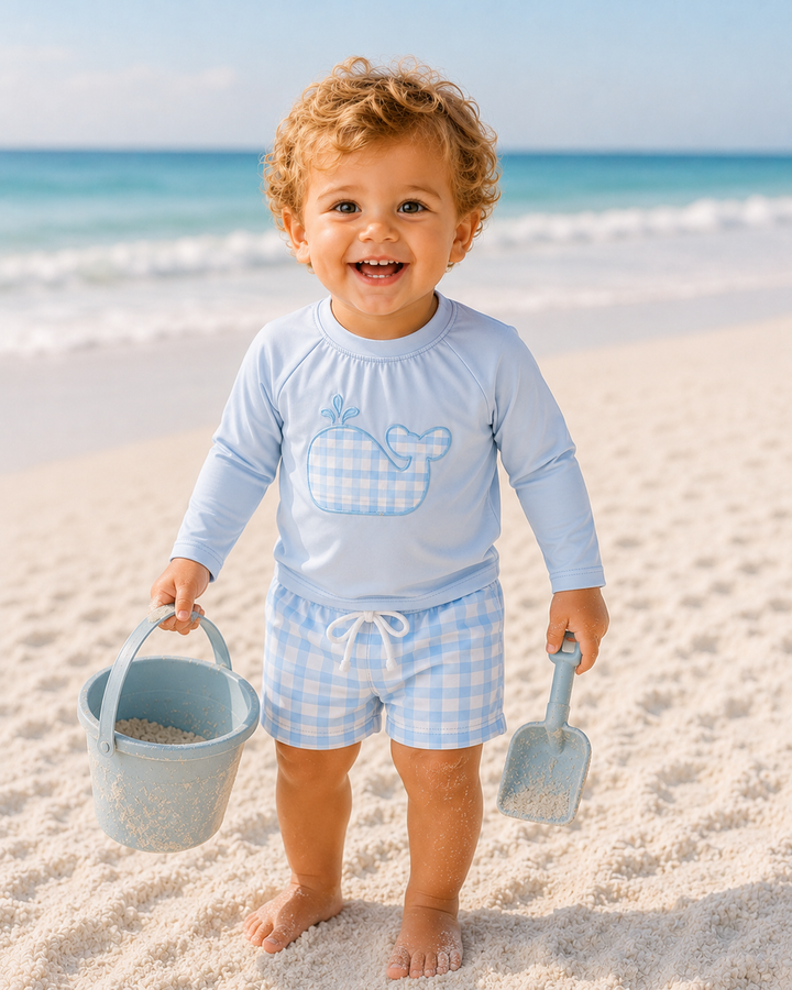 Child in light blue rash guard and checkered shorts playing on a sandy beach with ocean in the background