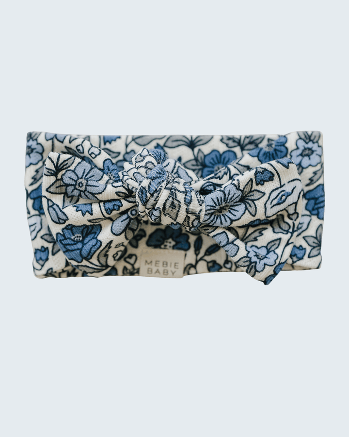 A fabric headband with a knotted bow detail, featuring a blue floral and leaf pattern on a cream background. The brand name &