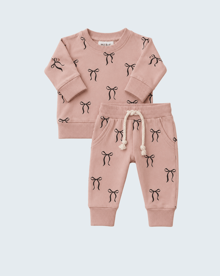 A two-piece baby clothing set in dusty pink, featuring a long-sleeve top and matching jogger pants. The set is decorated with a repeating black bow print, and the pants have a cream-colored drawstring.