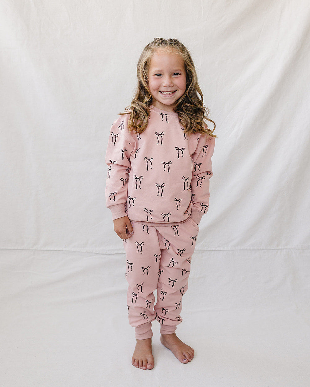 A young girl with blonde hair stands barefoot against a white fabric backdrop, wearing a matching dusty pink pajama set with a black and white bow pattern.