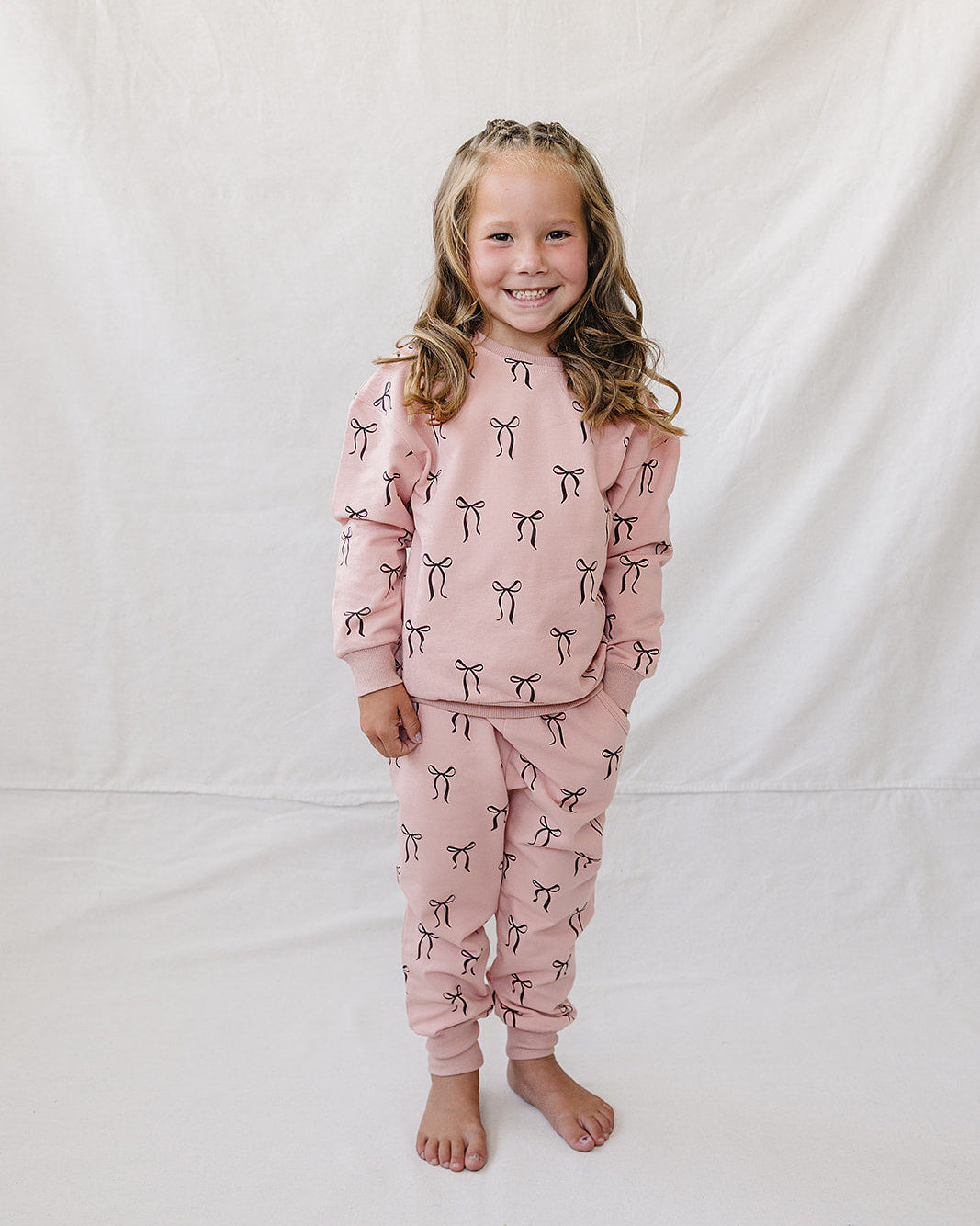 A young girl with blonde hair stands barefoot against a white fabric backdrop, wearing a matching dusty pink pajama set with a black and white bow pattern.