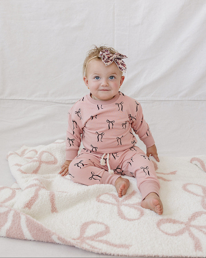 A baby sitting on a white blanket with pink bow patterns, wearing a matching pink long-sleeve romper and a pink bow headband.