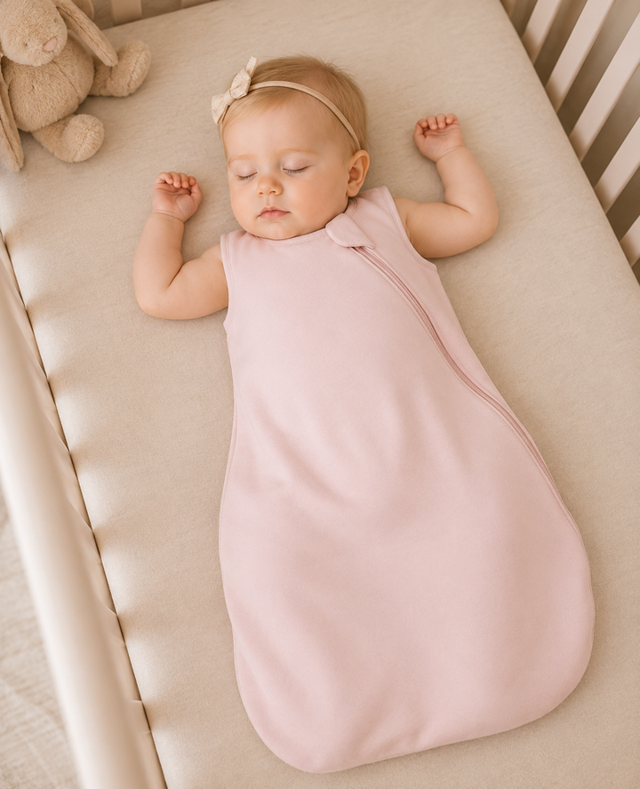 A sleeping baby lies on their back in a crib, swaddled in a pale pink sleep sack with a matching headband.