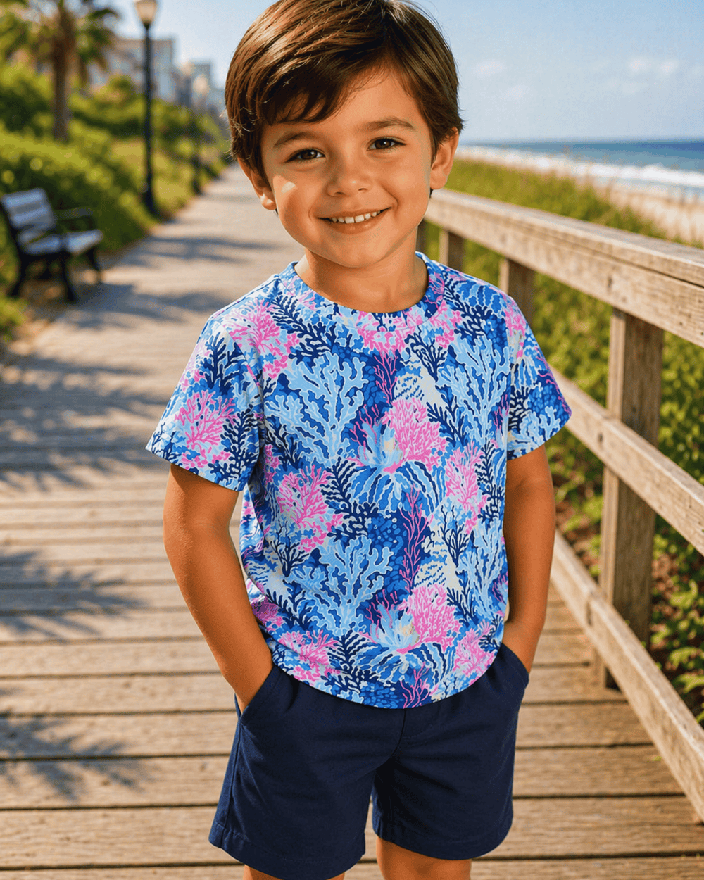 Child wearing a blue floral shirt and navy shorts standing on a wooden boardwalk by the beach.