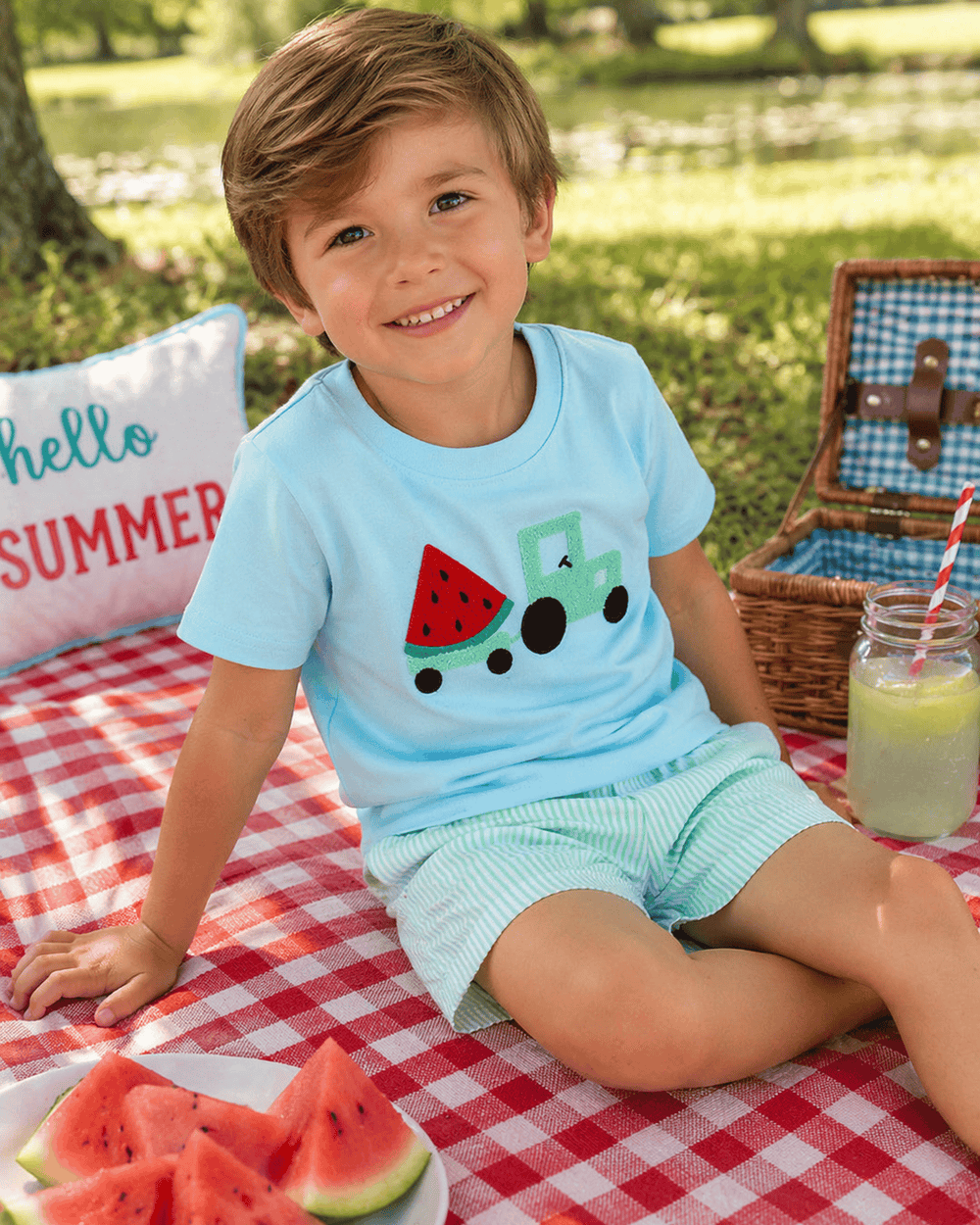Child sitting on a picnic blanket with watermelon slices and a lemonade drink, wearing a light blue shirt with a watermelon design.