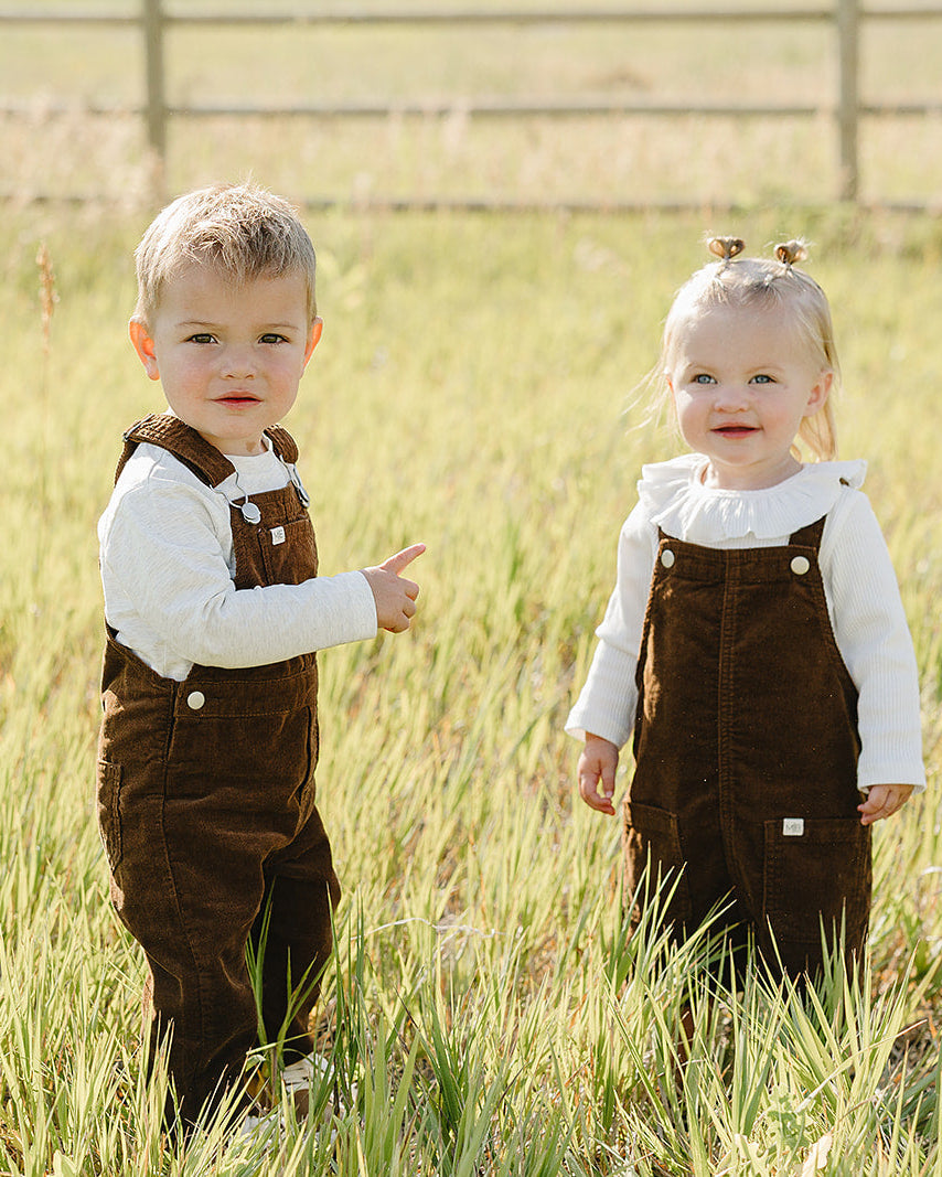 Brown corduroy overalls on two toddlers, adjustable straps and front patch pockets.