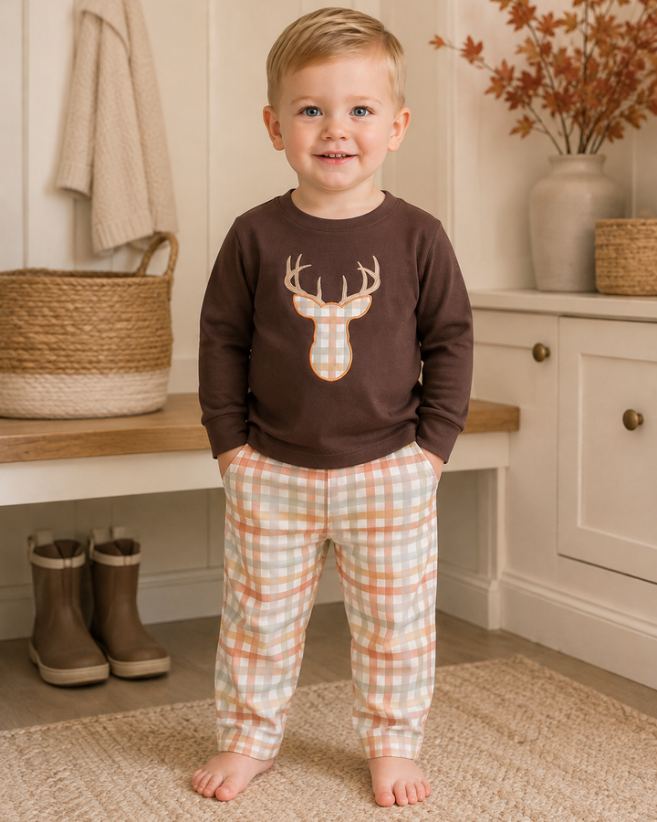 A young child standing barefoot in a cozy indoor room, wearing a dark brown long-sleeve shirt with a deer head graphic and peach-and-white plaid pants.
