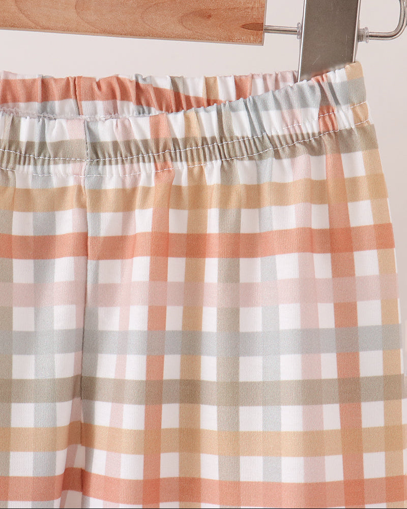 A close-up of a plaid pants hanging from a wooden hanger with a metal clip. The fabric has an elastic waistband and features a checkered pattern in white, peach, beige, and light gray.