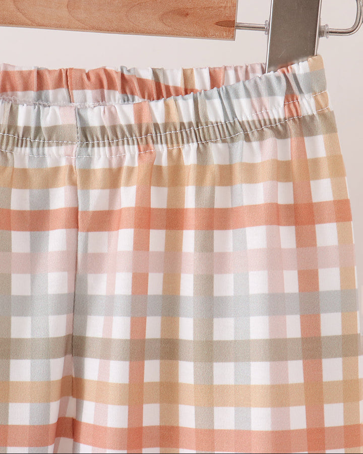 A close-up of a plaid pants hanging from a wooden hanger with a metal clip. The fabric has an elastic waistband and features a checkered pattern in white, peach, beige, and light gray.