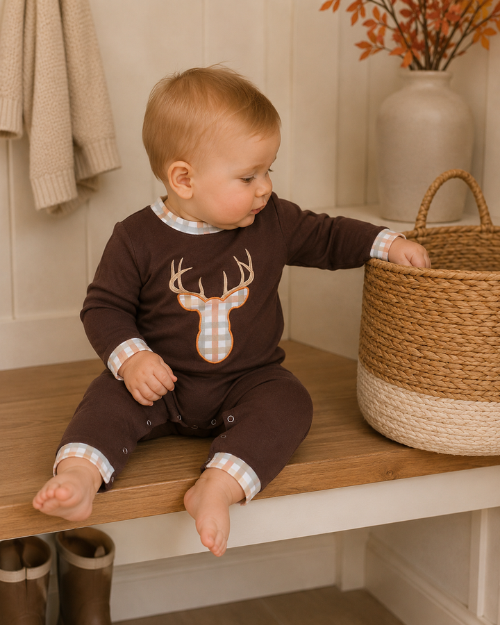 A baby sitting on a wooden bench wearing a dark brown long-sleeve romper with a deer graphic and plaid trim, next to a woven basket in a cozy indoor setting.