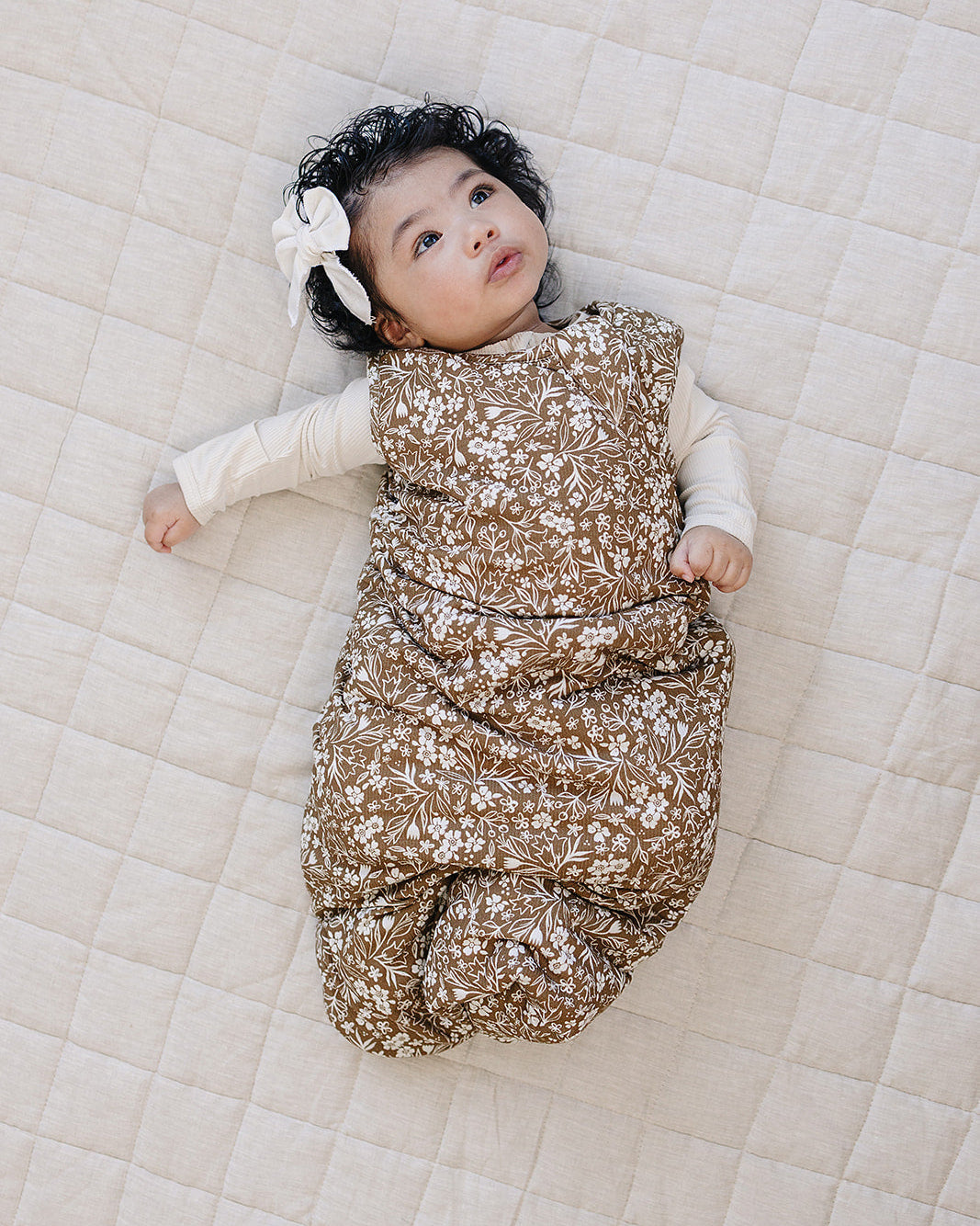 Baby lying on a quilted surface wearing a floral sleep sack and white headband.