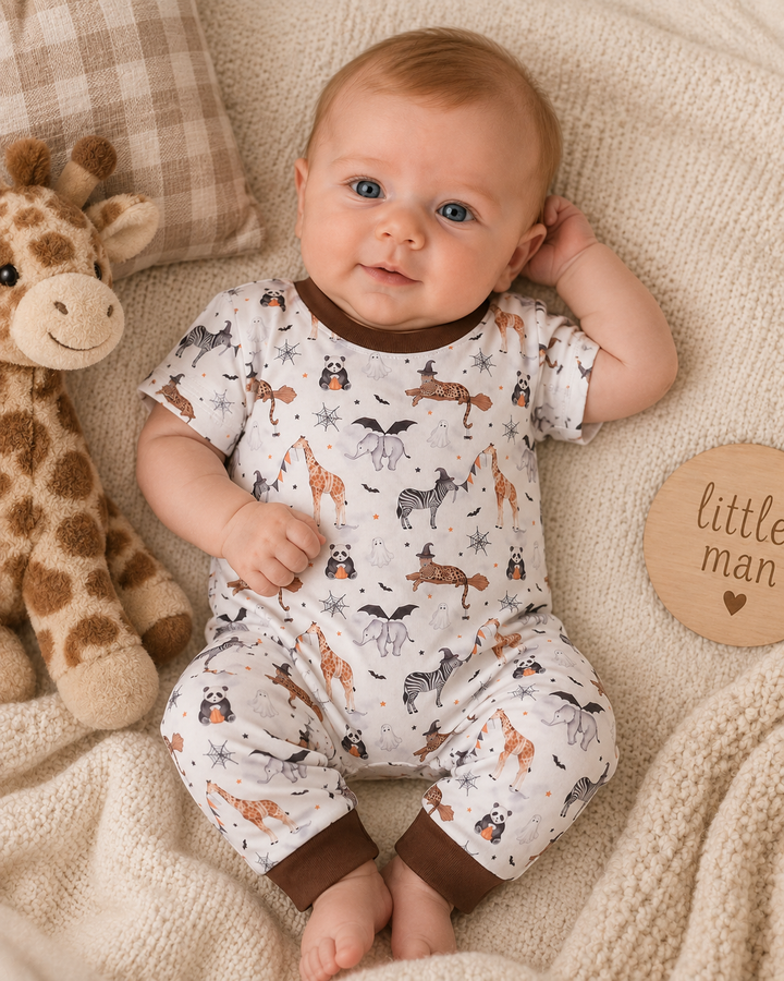 A baby lying on a cream knit blanket next to a plush giraffe toy, wearing a short-sleeve footless onesie with a woodland animal print and brown trim. A round wooden tag reading &