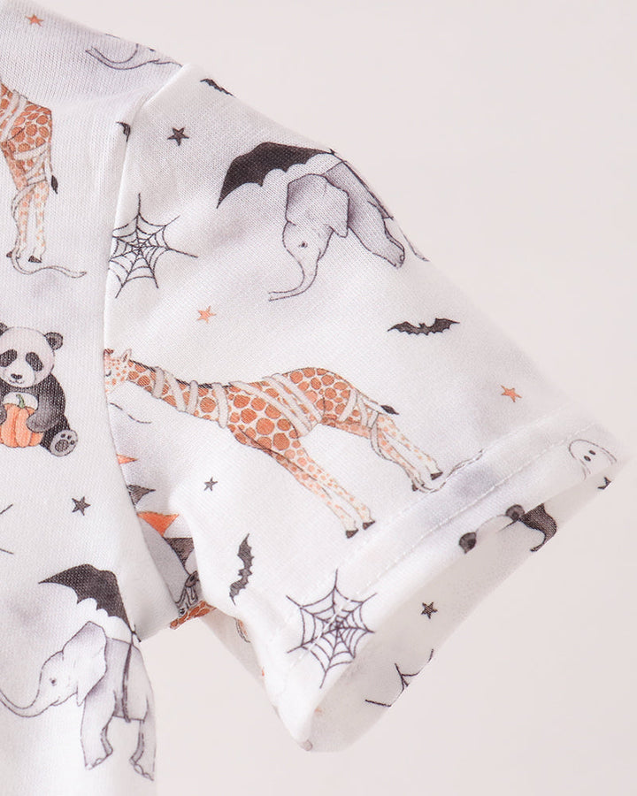 A close-up of a white baby garment featuring a playful print of orange-brown giraffes, black bats, gray elephants, pandas, and spiderwebs, set against a solid light beige background.