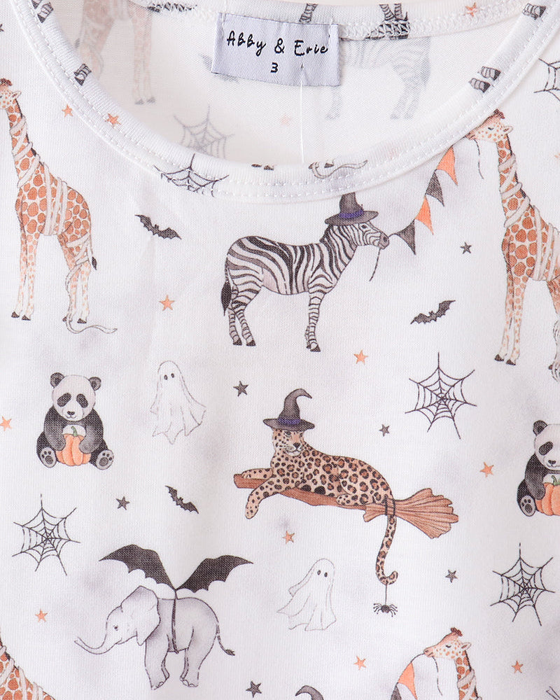 A close-up of a white baby top on a wooden hanger, featuring a colorful Halloween-themed animal print with a zebra, giraffe, panda, ghost, elephant, witch cat, spiderwebs, and bats. The brand label reads &