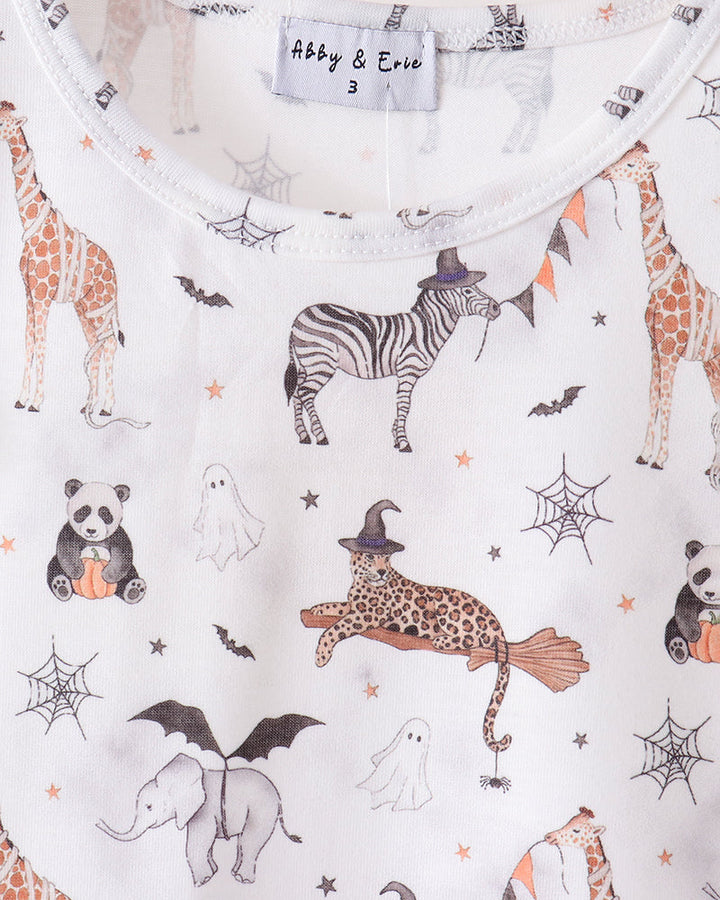 A close-up of a white baby top on a wooden hanger, featuring a colorful Halloween-themed animal print with a zebra, giraffe, panda, ghost, elephant, witch cat, spiderwebs, and bats. The brand label reads &