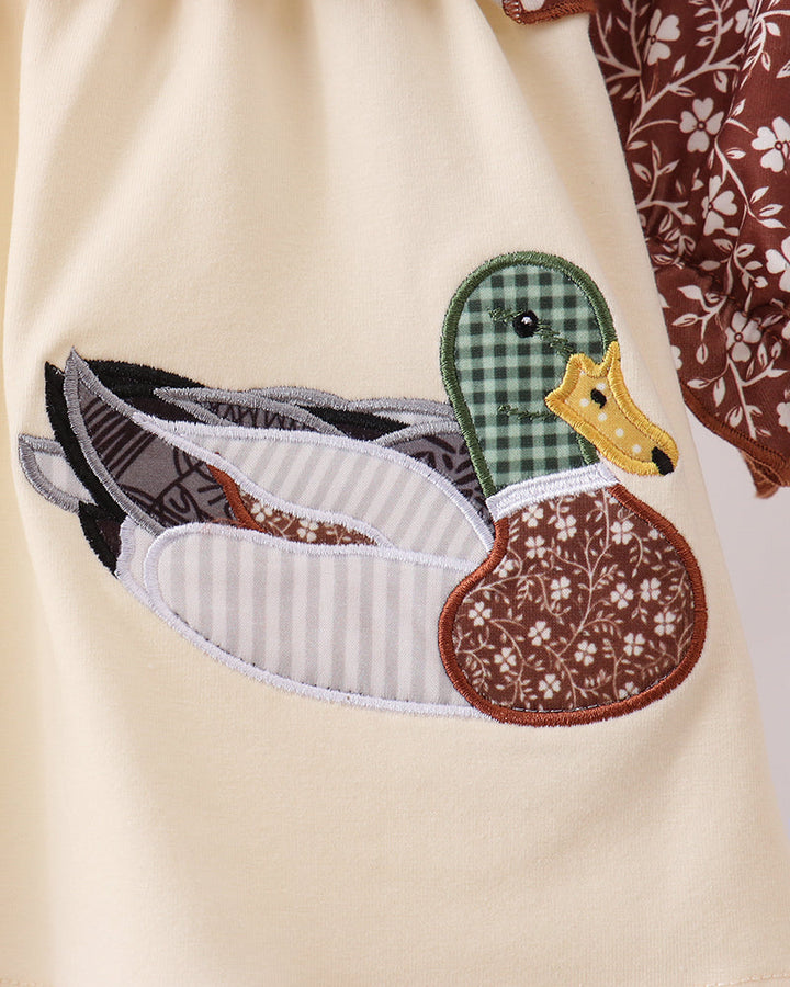 Close-up of a cream-colored garment featuring a detailed applique patch of a duck on the front. The patch includes a green gingham head, a yellow beak, a brown patterned body, and gray-and-white striped wings.