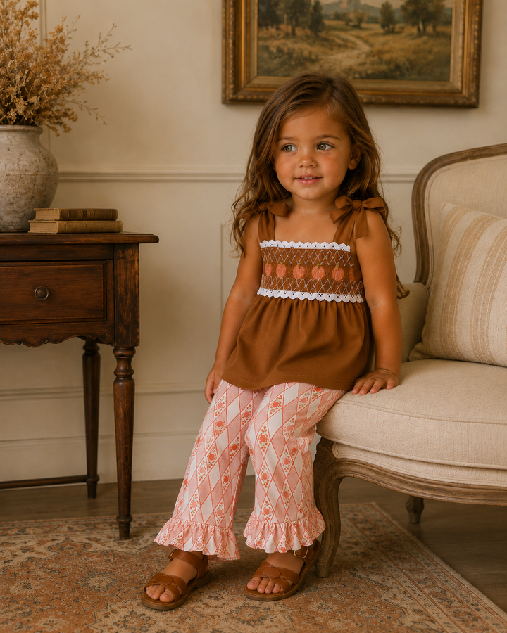 A young girl wearing a brown smocked sleeveless top with white lace trim and pink patterned ruffled pants, sitting on a beige armchair in a warm, traditional room.