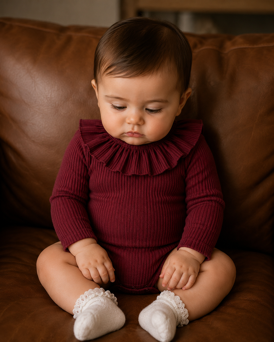 A baby sitting on a brown leather couch wearing a burgundy ribbed long-sleeve romper with a ruffled collar and white lace-trimmed socks.
