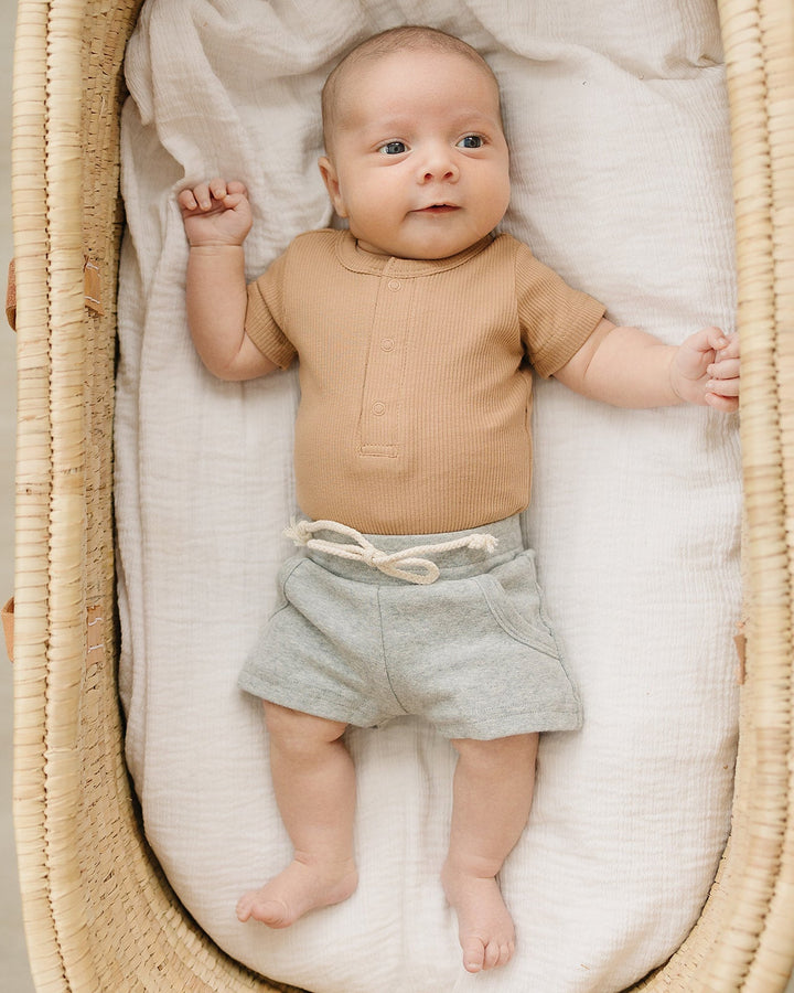 Baby lying in a moses basket wearing a brown shirt and gray shorts.