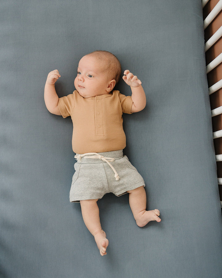 Baby lying on a gray surface wearing a brown shirt and gray shorts.