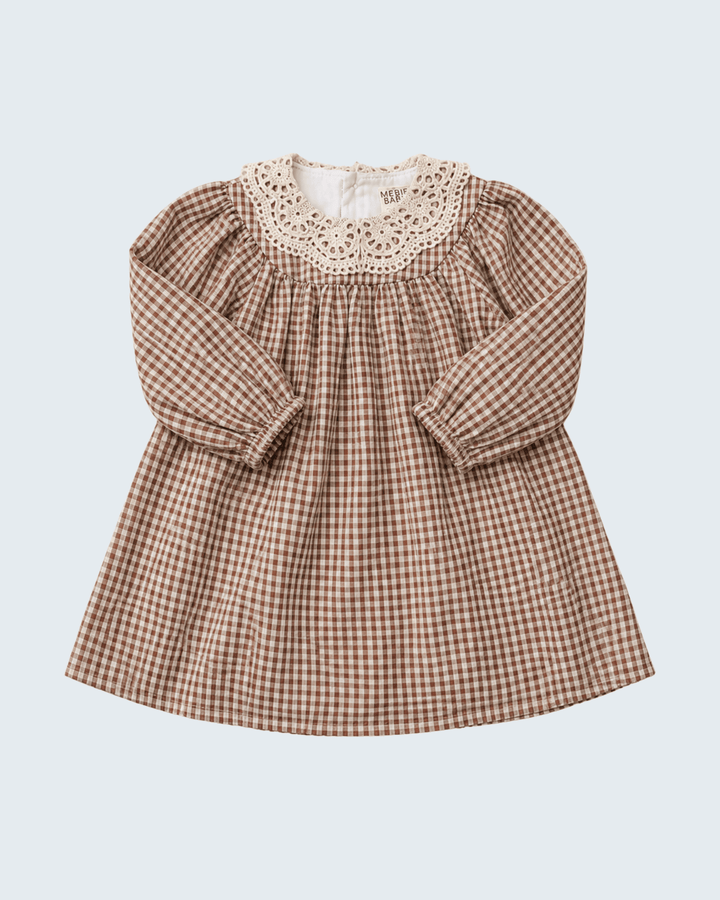 A long-sleeve baby dress with a brown and beige gingham pattern, featuring a smocked bodice and a cream-colored crochet lace collar, shown against a light gray background.