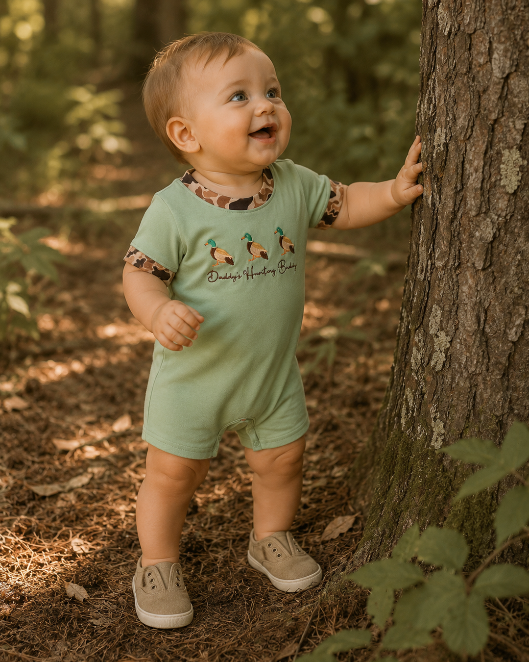 Baby in a green romper standing next to a tree in a forest
