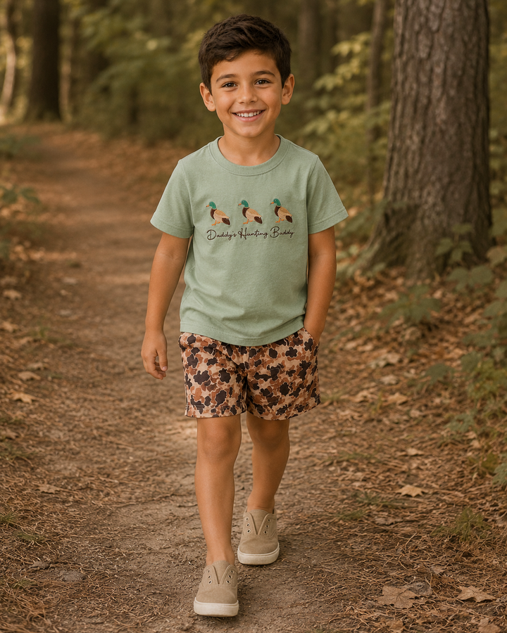 Child wearing a green t-shirt with duck design and floral shorts in a forest setting