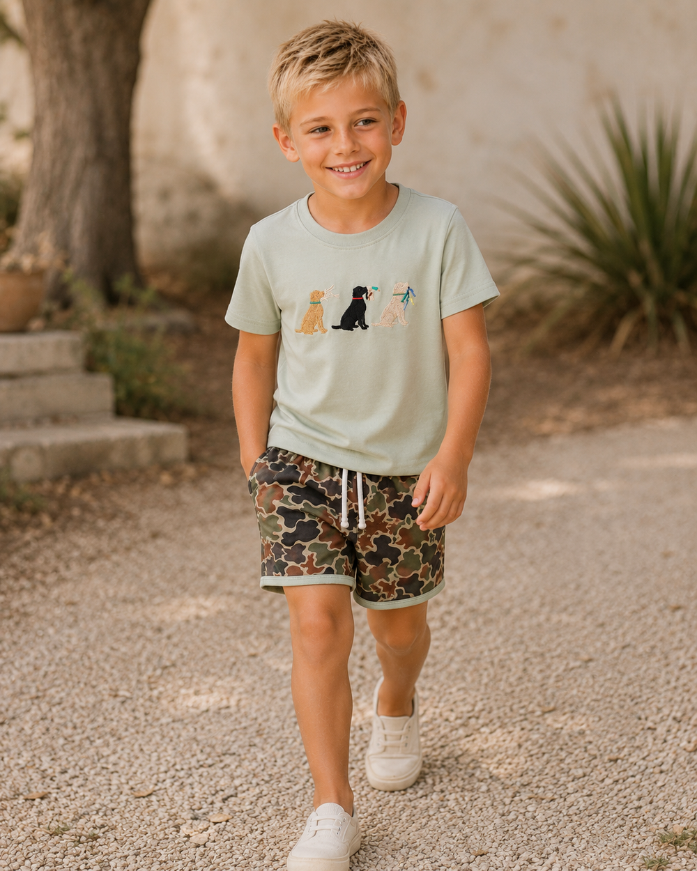 Young boy wearing a light green t-shirt with dog illustrations and camouflage shorts, standing outdoors.
