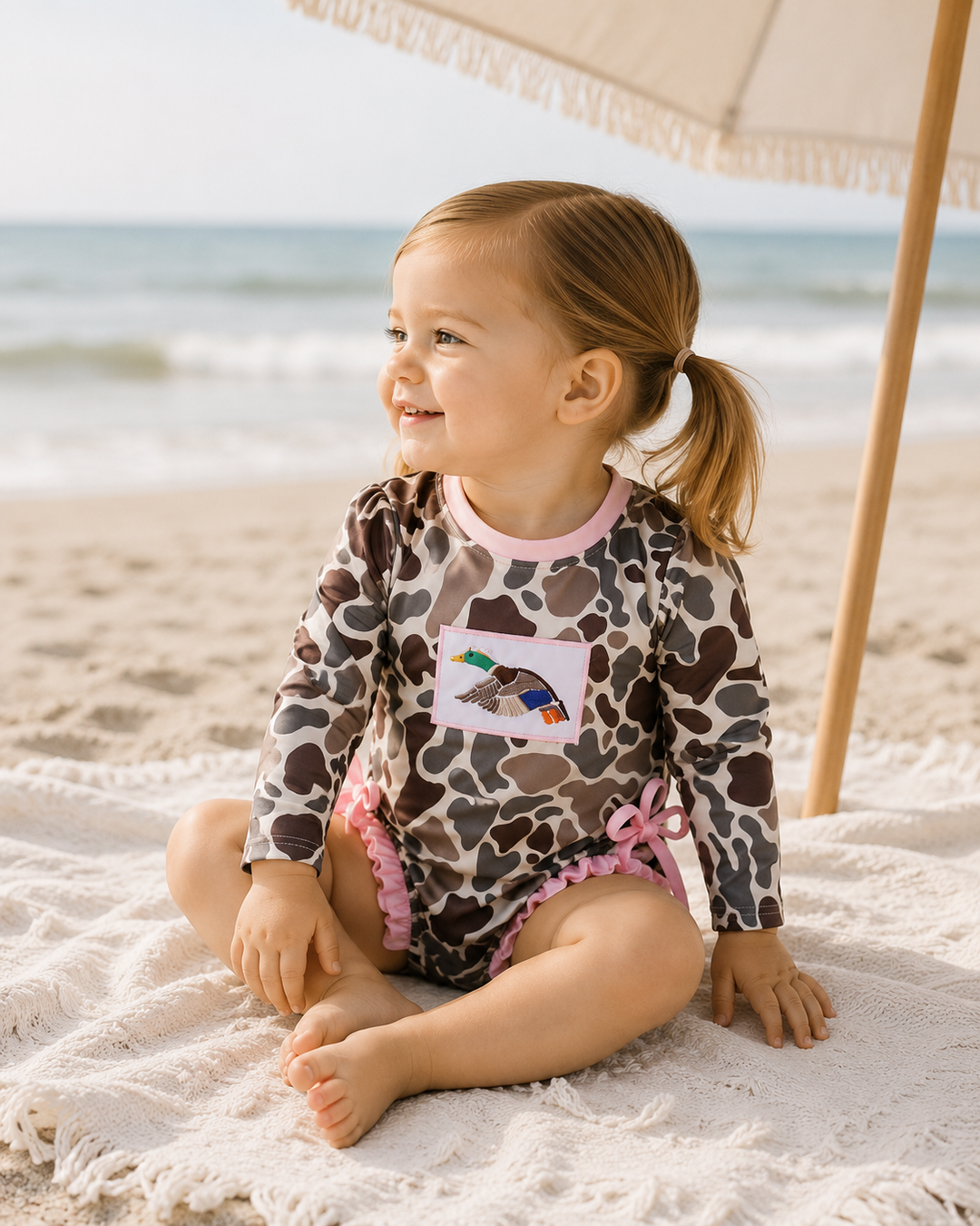 Child in a leopard print swimsuit sitting on a towel at the beach with an umbrella in the background.