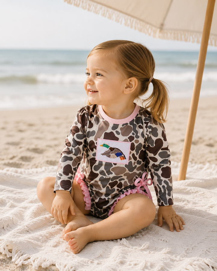 Child in a leopard print swimsuit sitting on a towel at the beach with an umbrella in the background.