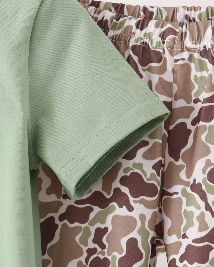 A close-up of a matching two-piece clothing set featuring a light sage green short-sleeve top and camouflage print shorts in shades of green, brown, and beige.