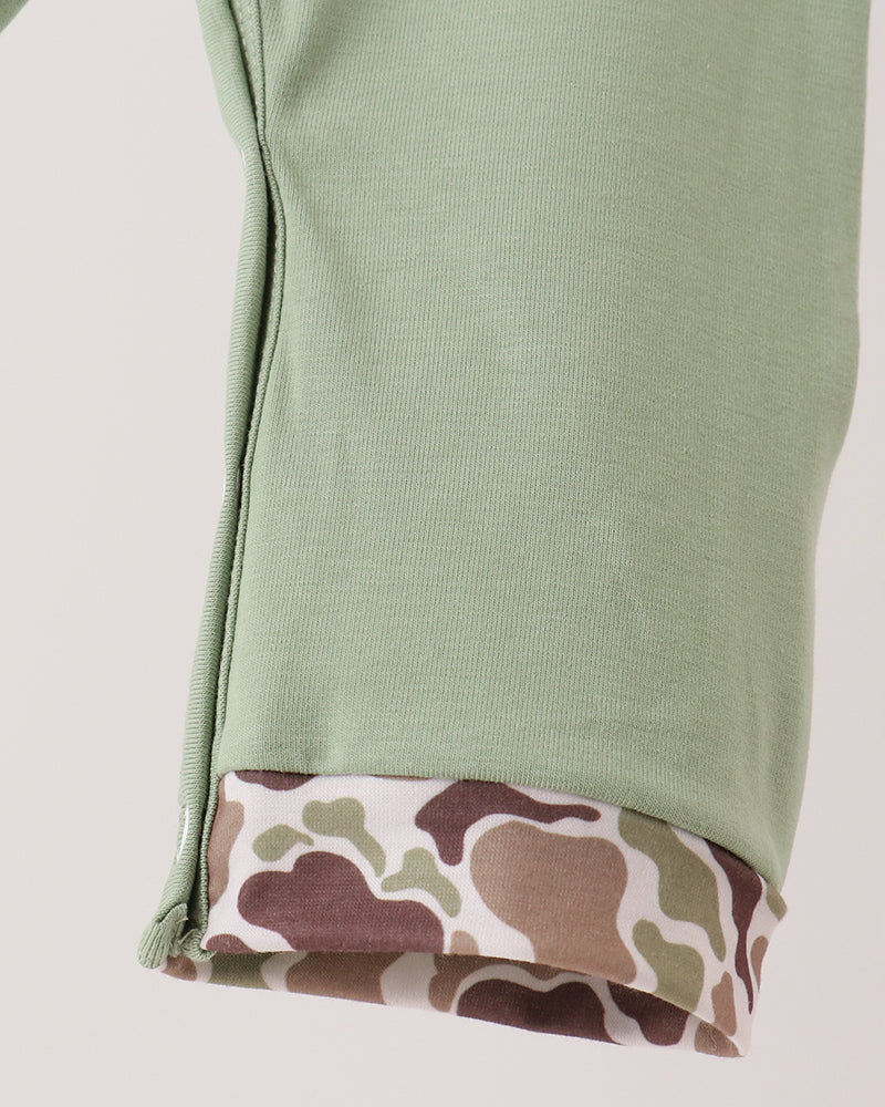 Close-up of a sage green ribbed knit garment cuff with a folded camouflage-patterned lining showing a zipper, on a light gray background.