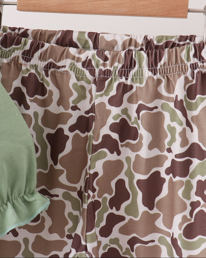 Camouflage print shorts in brown, beige, and green hanging on a wooden hanger, with a light green ruffled sleeve partially visible on the left.
