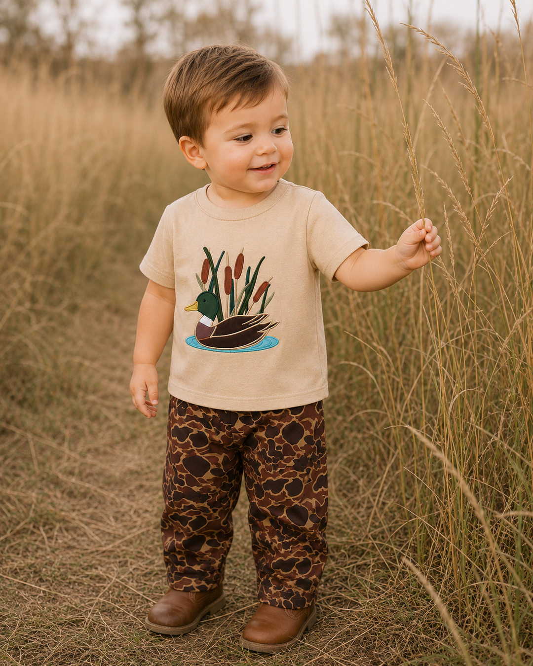 A toddler standing in a field of dry grass, wearing a beige t-shirt with a duck and cattails graphic, brown patterned pants, and brown shoes.