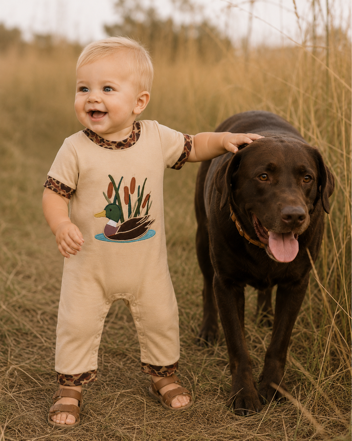 A baby wearing a beige one-piece romper with a duck graphic and leopard-print trim stands in a field next to a large brown dog.