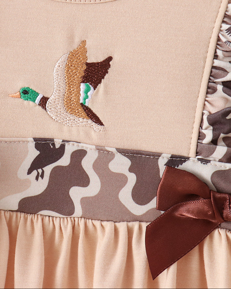 Close-up of a beige baby dress featuring an embroidered flying duck on the chest, a brown satin bow, and a camouflage patterned waistband.