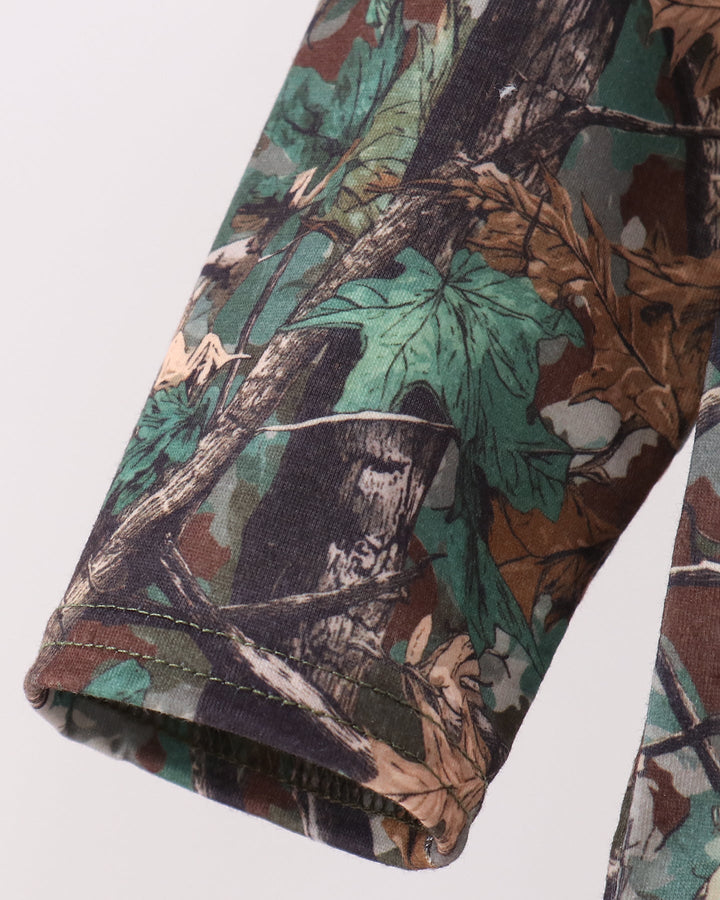 Close-up of a camouflage-patterned garment sleeve with a forest camo print featuring green, brown, black, and beige tones, shown against a plain light background.