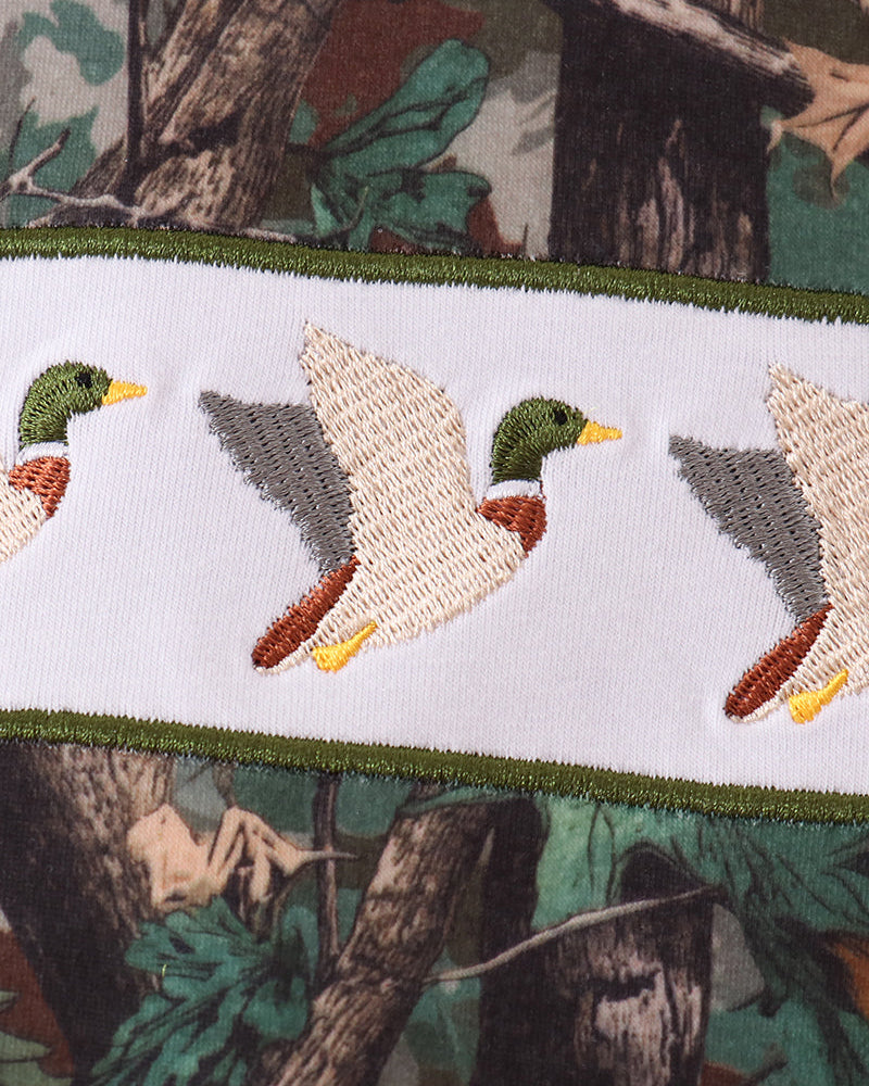 Close-up of a white embroidered band featuring repeating mallard duck motifs in green, yellow, brown, and gray, bordered by green trim and set against a woodland camouflage background.