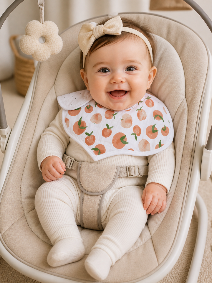 Baby in a beige baby seat wearing a bib with peach pattern and a bow headband.