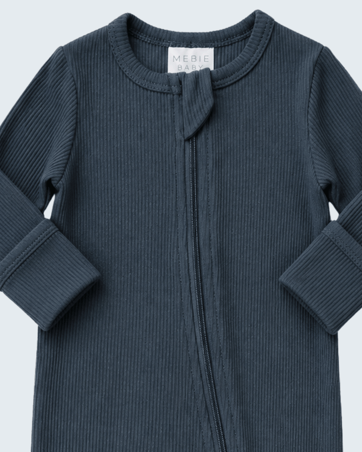 A close-up of a ribbed long-sleeve baby sleep sack or footed sleeper in a muted slate blue-gray color, featuring a front zipper and a small tie detail at the neckline. The brand label &