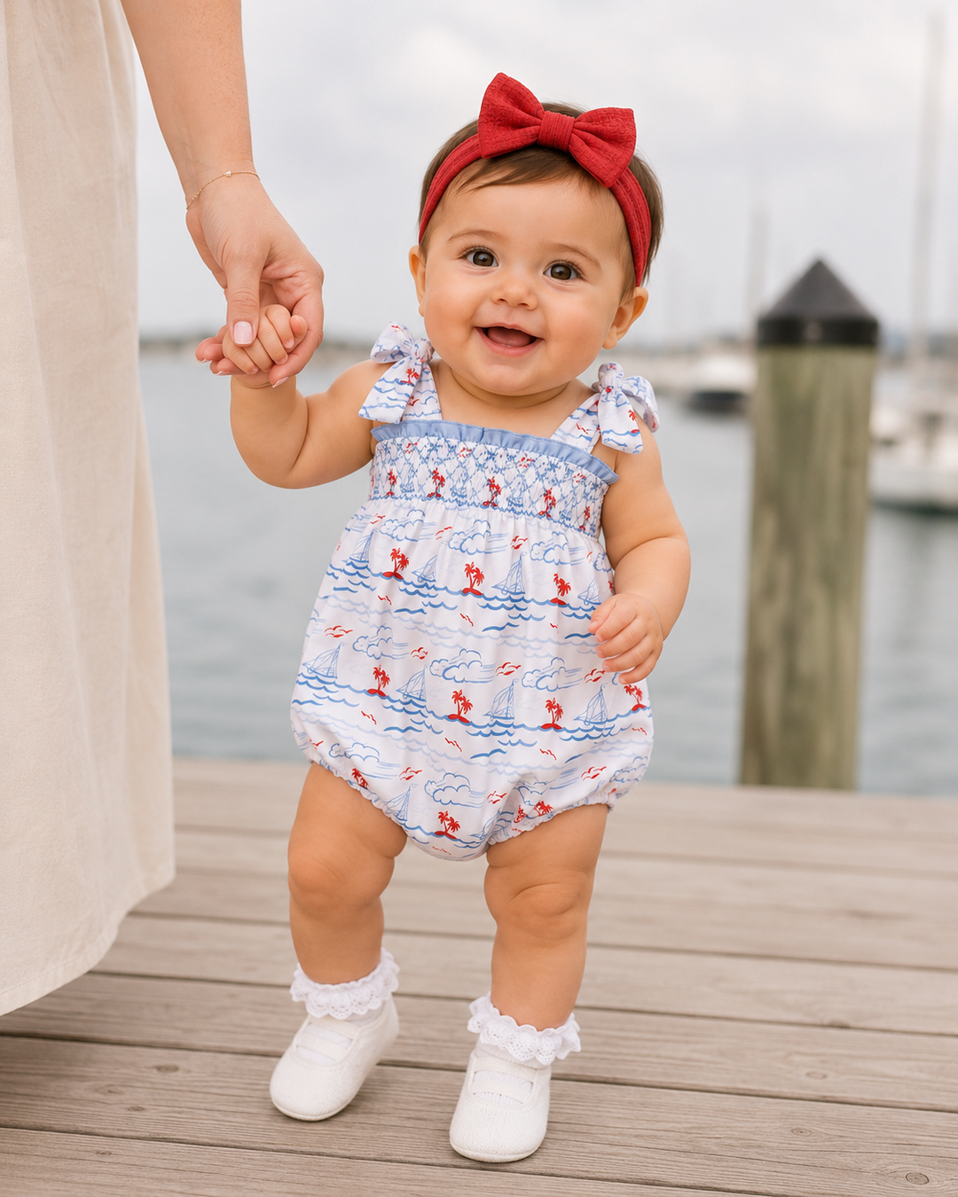 Baby in a floral romper with a red bow standing on a dock by water.