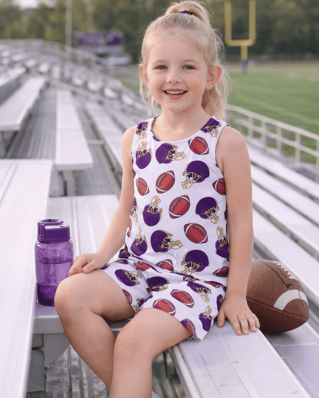 Young girl sitting on bleachers wearing an outfit with sports-themed print, with a football and water bottle nearby.